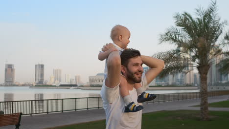 loving father in a white t-shirt in the summer walks with a child sitting on the neck against the city and palm trees