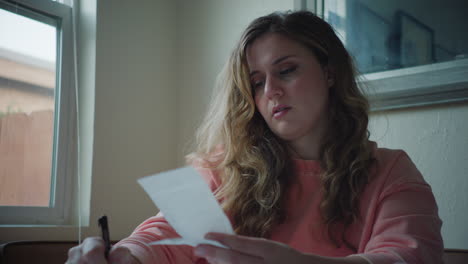 woman in pink sweater stressed out by bills and receipts at kitchen table in daytime, sitting by window