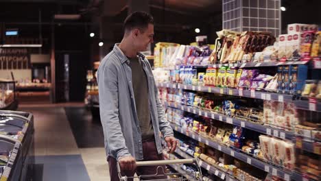 Im-Supermarkt:-Glücklicher,-Stylischer-Mann-Mit-Einkaufswagen-In-Einem-Großen-Einkaufszentrum,-Der-An-Den-Regalen-Mit-Waren-Vorbeigeht-Und-Plötzlich