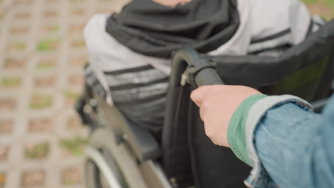 Parent Pushing Child In Wheelchair With Green Cap, Closeup Of Hand On Handle And Wheel, Compassionate Outdoor Outing, Denim Jacket And Casual Shoes Visible, Warm Family Support Atmosphere
