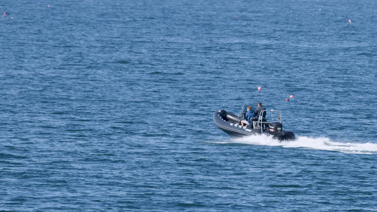 Two men ride a fast-moving rigid inflatable boat over calm blue sea under bright daylight, captured in a steady, wide shot with minimal camera movement