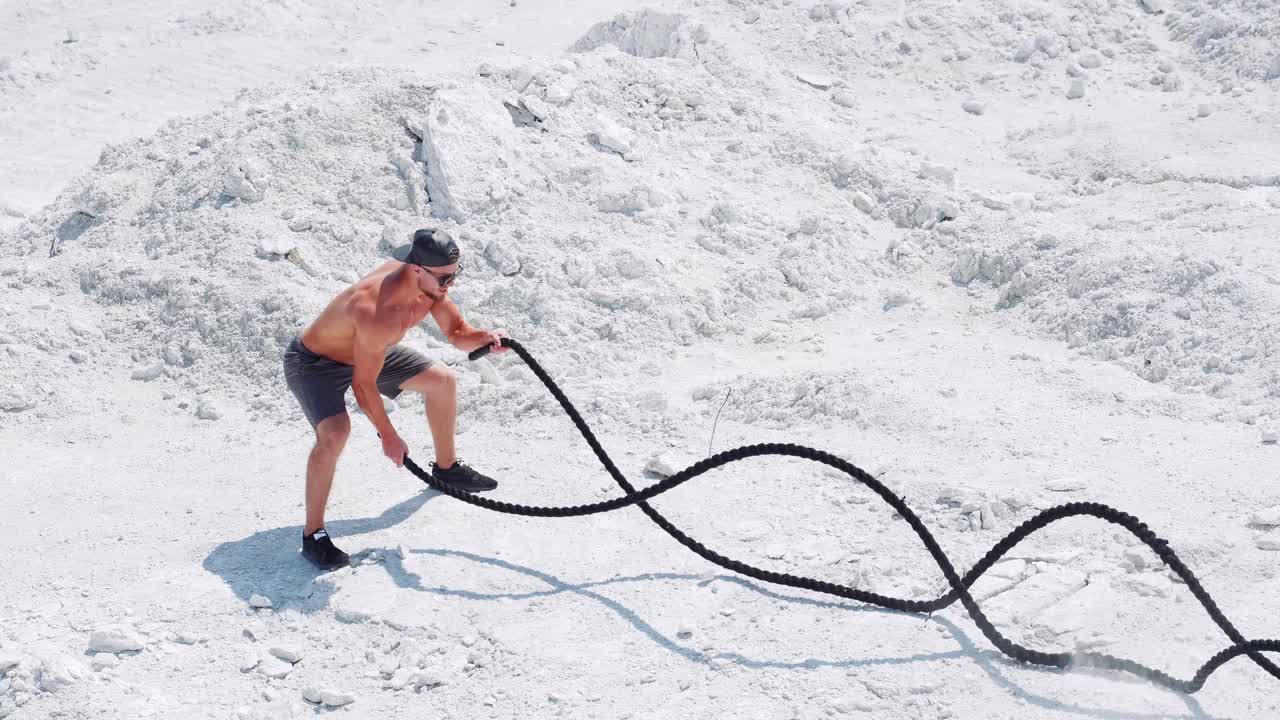 Sports activities with ropes on the street. Male athlete on the white sand does exercises with a rope