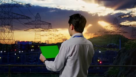 back view of an asian male professional worker standing near high voltage tower, industrial facility, checking on tablet with green screen and look at the factory once