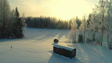 Aerial-drone-forward-moving-shot-over-rectangular-cottage-and-barrel-sauna-cabin-on-a-snow-covered-white-landscape-at-daytime