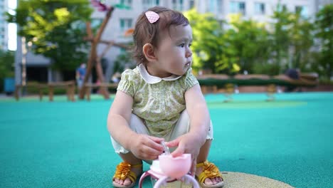 beautiful one-year-old little girl squatting in the playground playing with water bottle looking aside, looks lonely and sad because other kids refuse to play with her