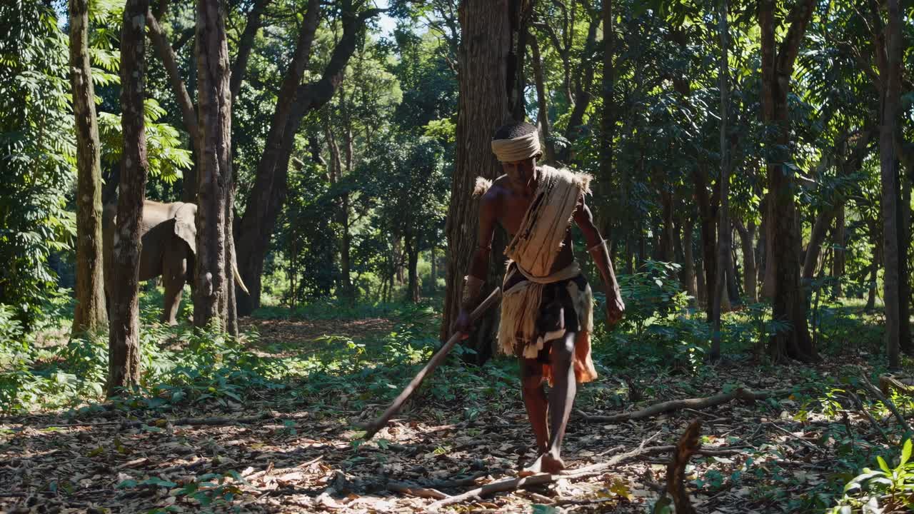 A man in traditional attire walks through a lush forest, captured from a low-angle shot