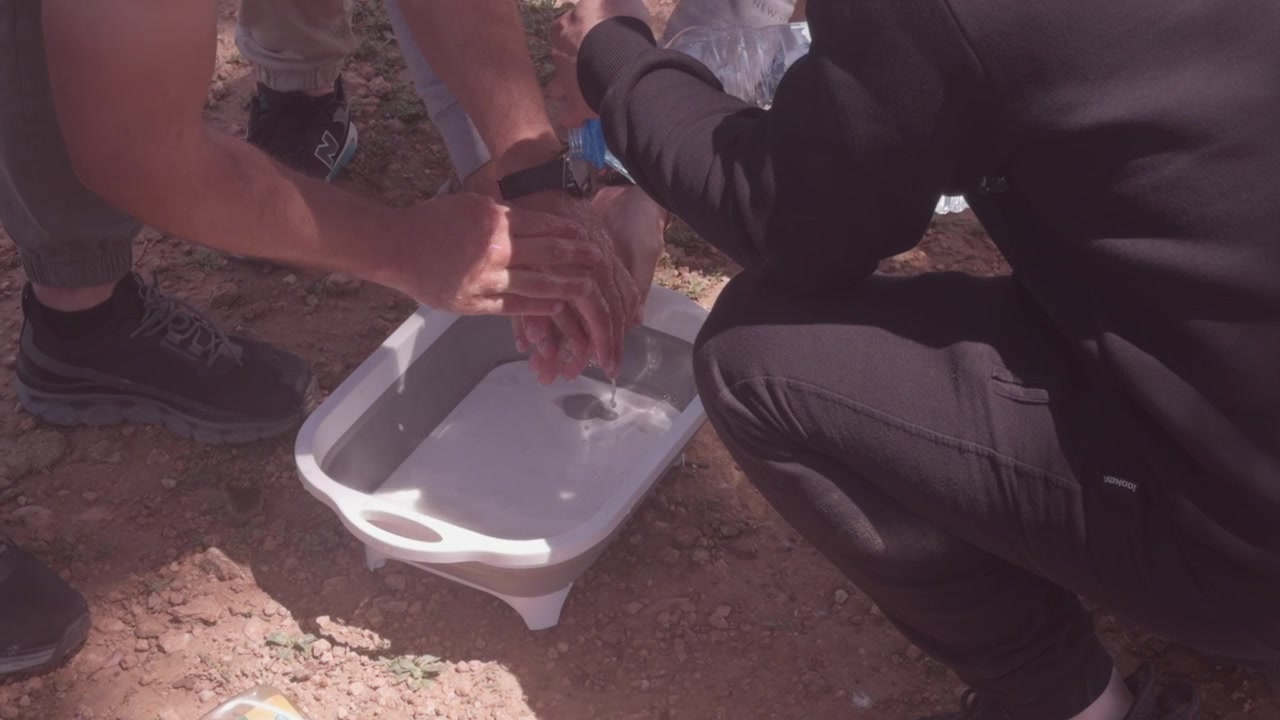 Persona che versa acqua per lavarsi le mani sopra una bacinella di plastica bianca su terreno roccioso e asciutto durante un picnic all'aperto in Marocco