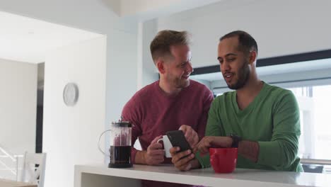 Multi-ethnic-gay-male-couple-in-kitchen-talking-one-using-smartphone
