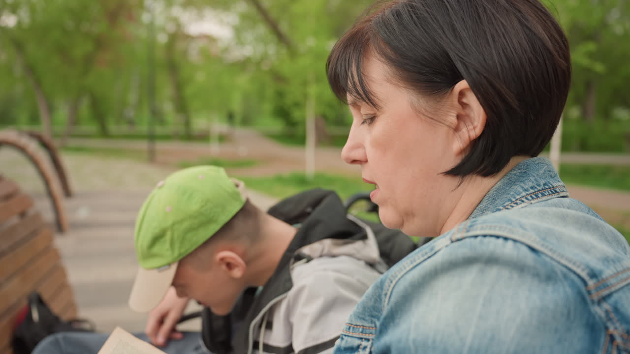 White Woman Reading Book With Boy, Park Bench Beside Wheelchair, Denim Jacket And Green Cap Visible, Engaged Expressions And Close Attention, Gentle Sunlight, Inclusive Learning And Bonding