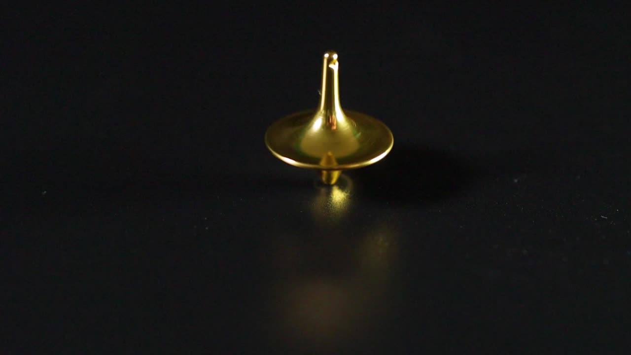 Um pequeno pião dourado gira constantemente sobre uma superfície preta lisa com reflexos sutis.