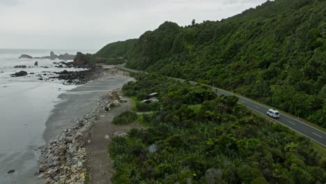 Stunning-aerial-view-of-tourist-campervan-on-a-road-trip-traveling-along-scenic-coastal-road-with-ocean-and-green-covered-landscape-on-the-West-Coast-of-South-Island,-New-Zealand-Aotearoa