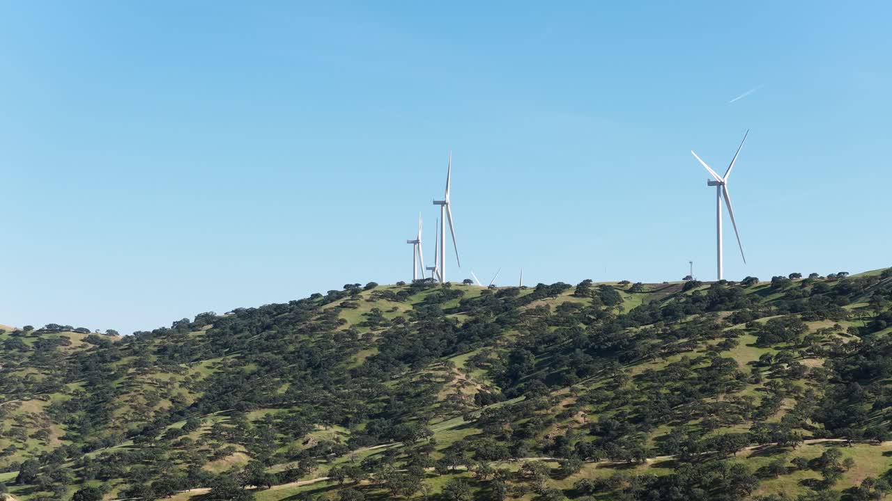 カリフォルニアのダイナソーポイント風力発電所で、豊かな緑の丘陵に立つ現代的な風力発電機が、澄み切った青空を背景に羽根を回し、クリーンな再生可能電力を生み出している。