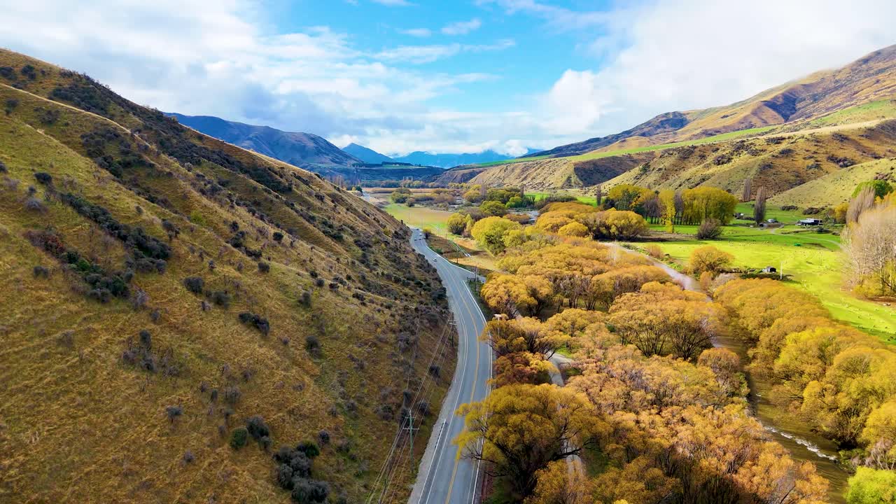 Drone glides above a winding rural road through a scenic valley with golden autumn trees, green farmland, and distant mountains under bright daylight