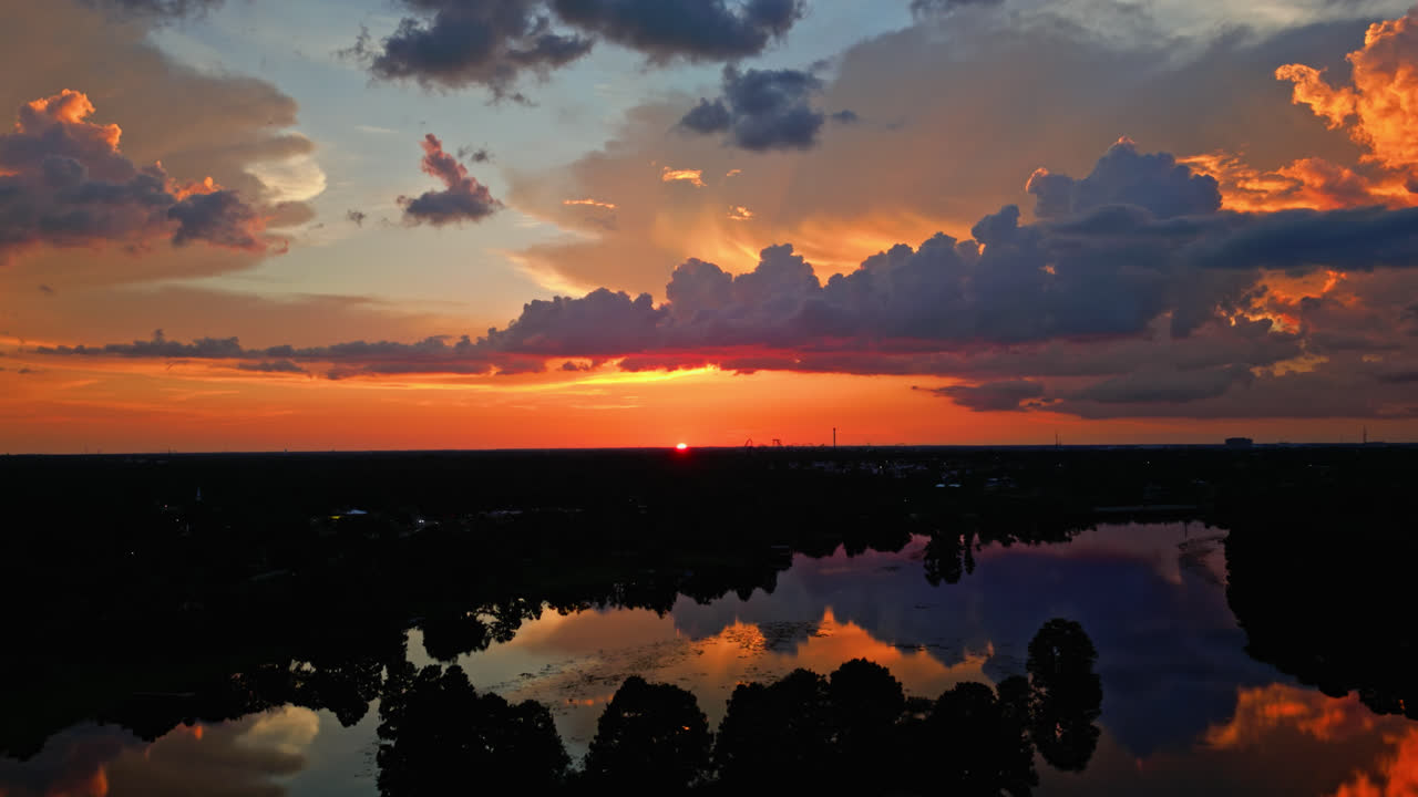 Sunset clouds reflect on still lake, soft orange and pink tones create tranquil backdrop as sun dips below horizon