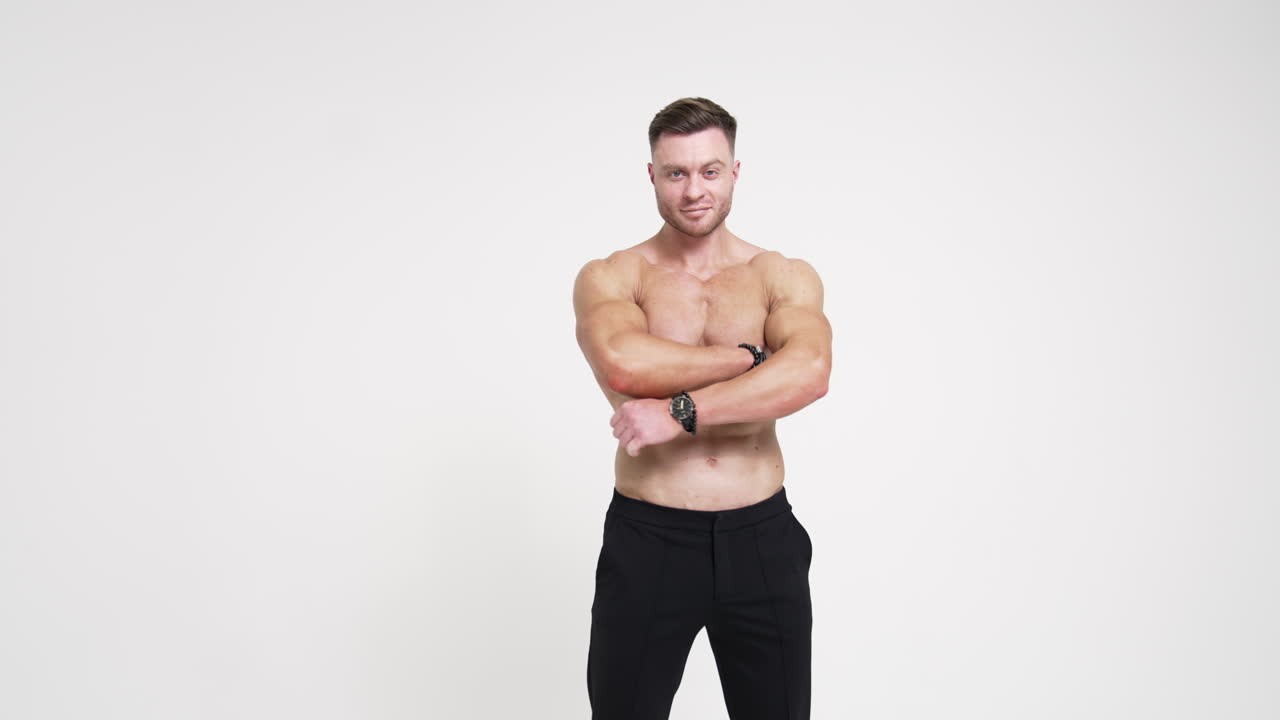Sportive man with naked torso holding hands in pockets of black trousers. Man crosses hands on chest smiling to camera. White backdrop.