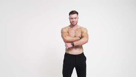 sportive man with naked torso holding hands in pockets of black trousers. man crosses hands on chest smiling to camera. white backdrop.