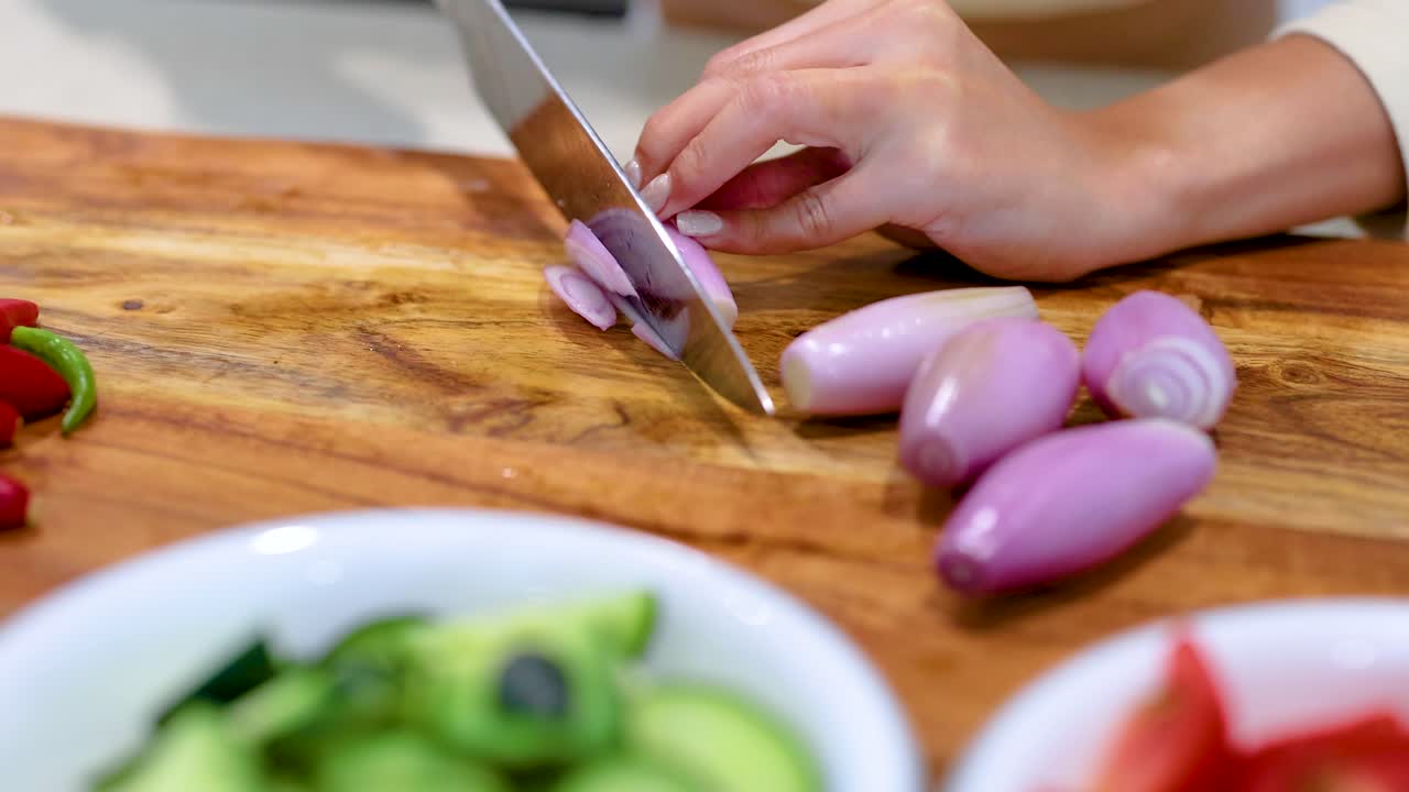 A hand slices shallots on a wooden board, surrounded by fresh vegetables. Bright lighting enhances the vibrant colors