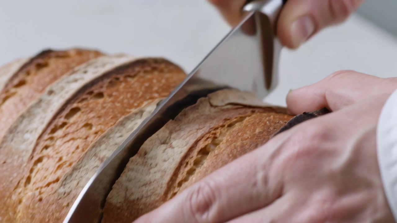 Slicing artisan loaf on counter, adult hands wearing chef shirt sawing after knife contacting crust