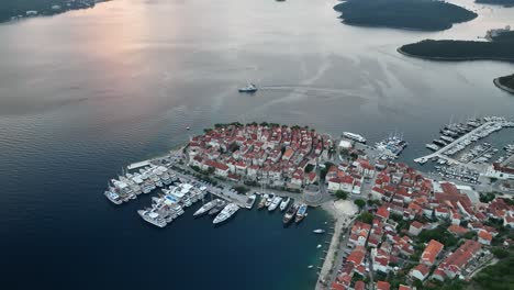 aerial drone video flying backwards from the old croatian town of korcula as a ferry is leaving, revealing the sunrise and surrounding mountains and islands