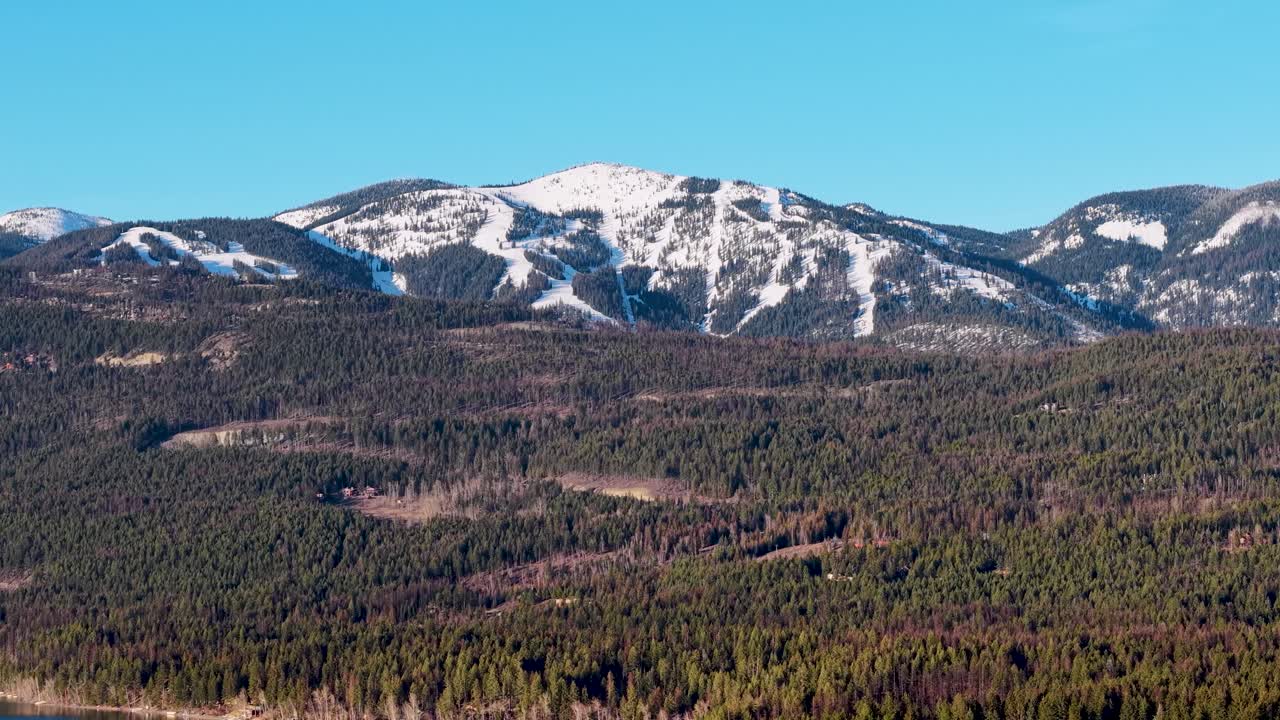 Luftfoto af en skovklædt bjergskråning, der fører til snelklædte tinder under en klar blå himmel med glimtende vand i det fjerne ved foden