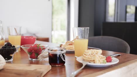 panqueques de desayuno en platos, con miel, fruta y jugo en la mesa en la cocina soleada, cámara lenta