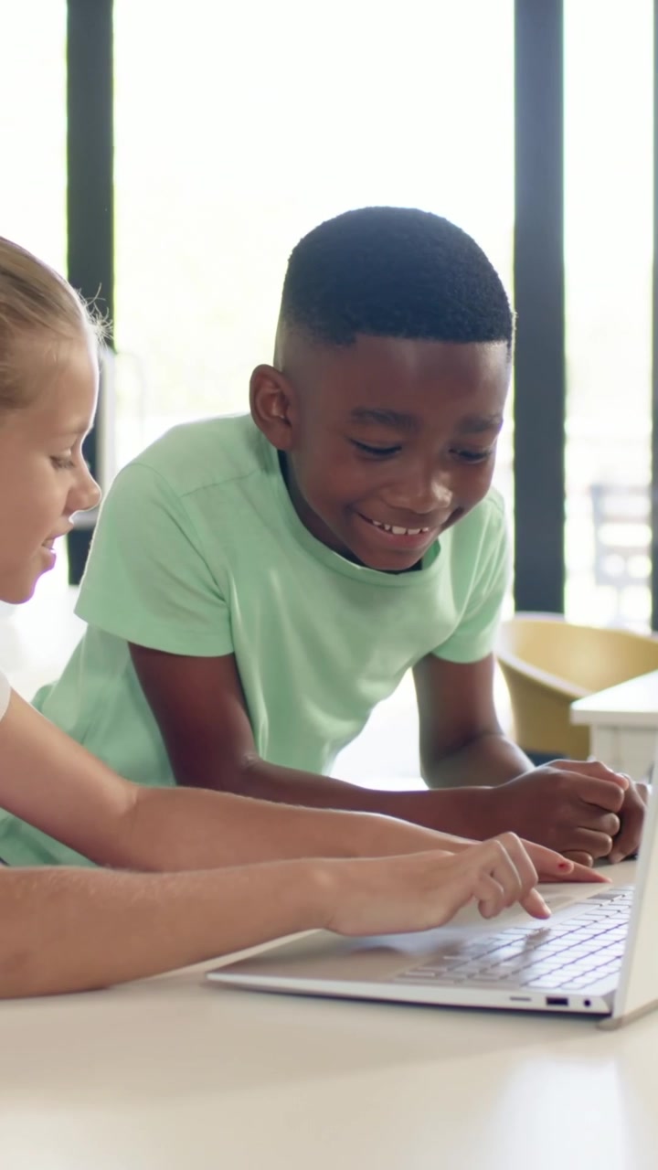 Vertical video: Leaning boy green tee watching girl white tee typing silver laptop at desk working
