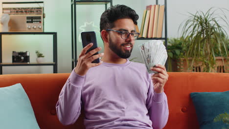 Smiling-happy-indian-man-counting-money-cash-use-smartphone,-income-saves-lottery-win-budget-at-home