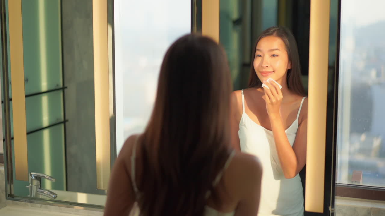 Beautiful Asian woman looking in mirror wiping face removing makeup. Pretty young lady in white dress applying cosmetics and using skincare routine. Young woman getting ready for a date in bathroom