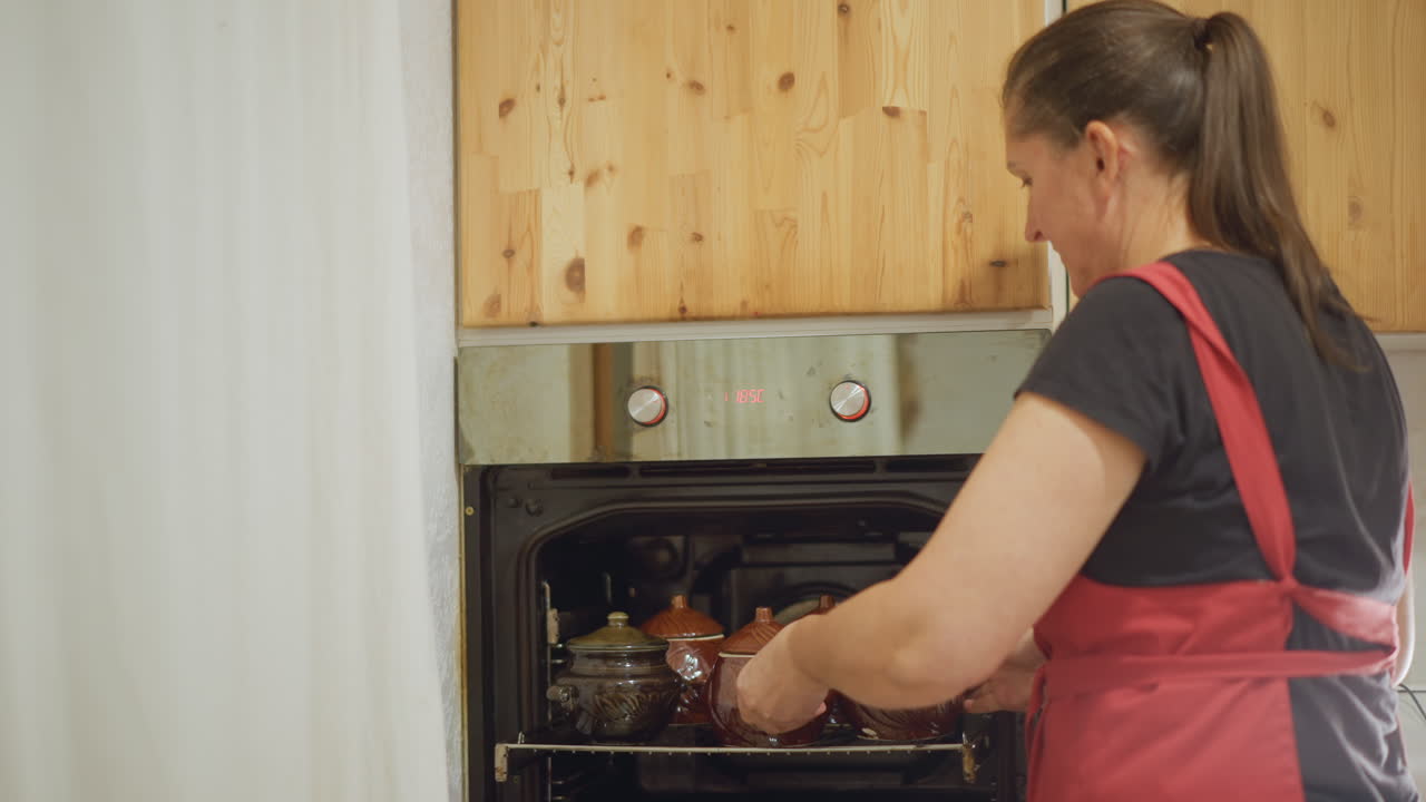 Donna bianca cuoca casalinga che mette pentole di ceramica nel forno, ambiente cucina rustica, grembiule rosso, mani attente che sistemano una casseruola di ceramica su un vassoio, mobili in legno, aroma che sale, calda atmosfera domestica