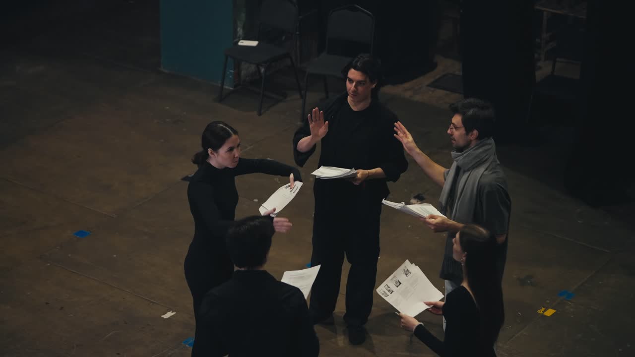 검은색 의상을 입은 자신감 넘치는 배우들이 원으로 서서 서로 대화를 나누고, 손에 스크립트를 든 종이를 들고, 극장 무대에서 연극과 공연에 대해 감독과 함께 이야기하고 있는 부분.