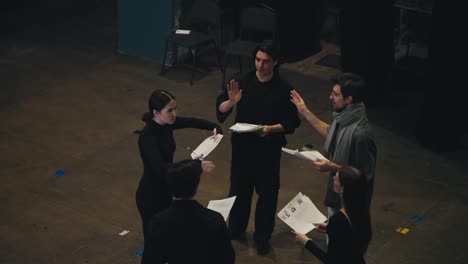 Top-view-of-a-group-of-confident-actors-in-black-suits-Standing-in-a-circle-and-talking-to-each-other-holding-sheets-of-paper-with-a-script-in-their-hands-and-talking-together-with-their-director-about-the-play-and-performance-on-stage-in-the-theater