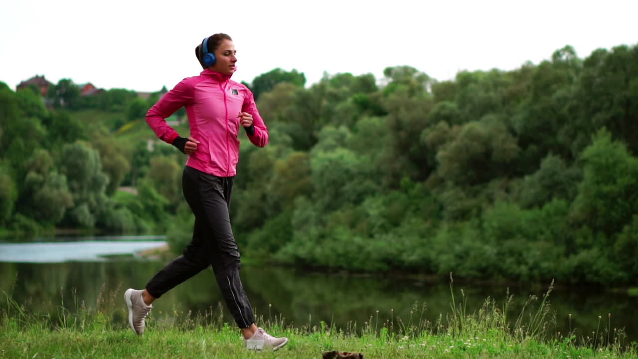 Una ragazza con una giacca rosa e pantaloni neri corre vicino al fiume, con le cuffie, preparandosi per la maratona.