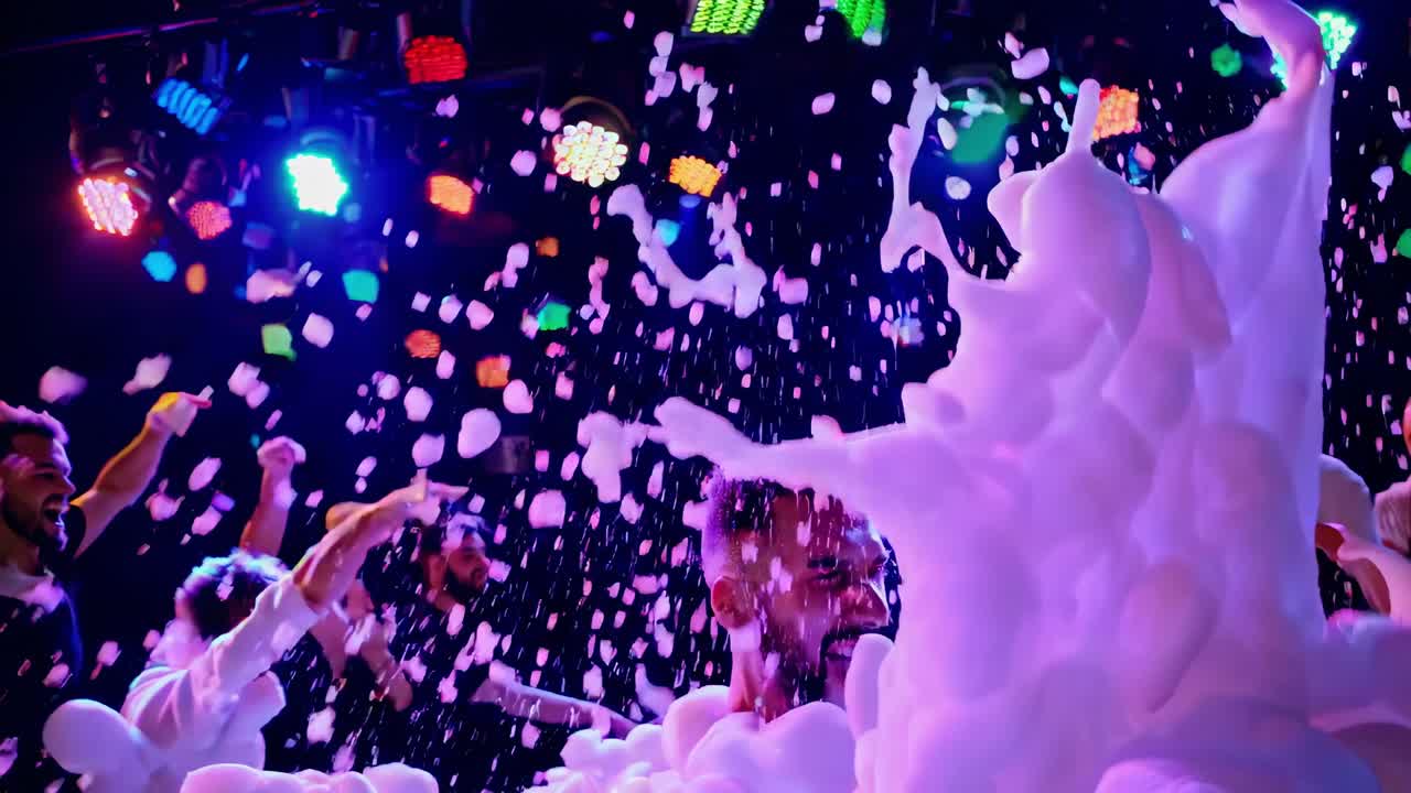 Young party crowd dancing enthusiastically, covered in foam and illuminated by vibrant stage lights, creating electrifying nightclub atmosphere with pulsing energy and collective joy