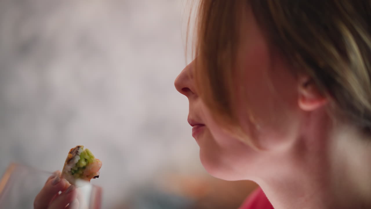 Young lady savoring sushi bite with chopstick as she enjoys taste in calm indoor space partially hidden behind hair with soft light and blurred background visible wine glass