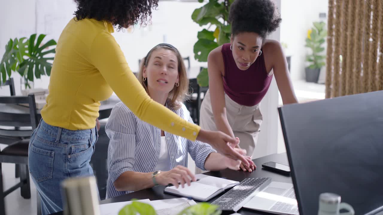 Diverse female team leaning in after seated woman gesturing, typing at large monitor to review work