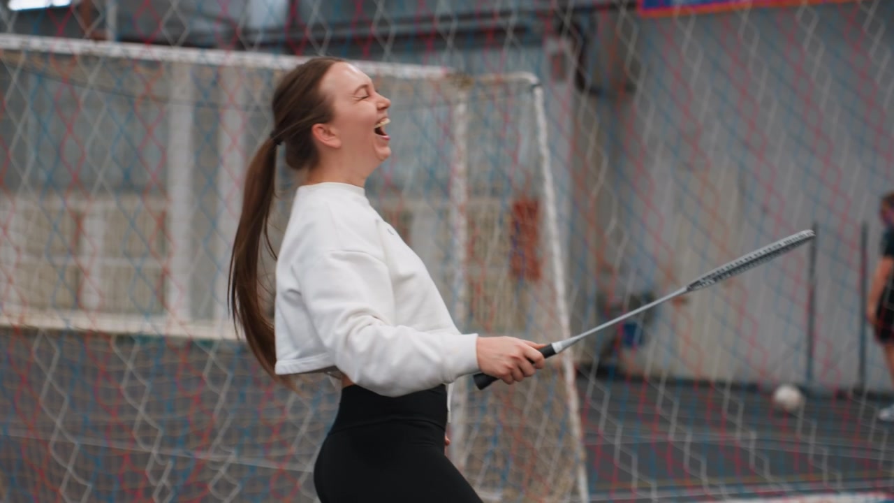 Young caucasian woman playing badminton in indoor court, laughing between rallies, casual training session with net backdrop, teammate jogging background, cozy sweatshirt, playful warmup, shuttlecock