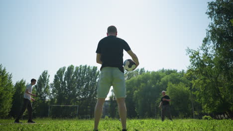 back view of a man rotating a soccer ball around him while the grandfather and his grandson plays football on a walk path close to a grassy field