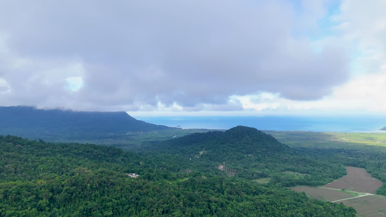 Drone camera slowly pans over dense rainforest and agricultural fields near the coast, revealing dramatic clouds, rolling hills, and distant ocean under soft daylight