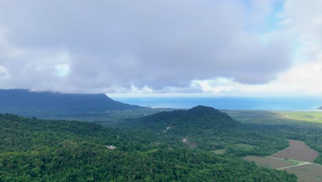 drone camera slowly pans over dense rainforest and agricultural fields near the coast, revealing dramatic clouds, rolling hills, and distant ocean under soft daylight