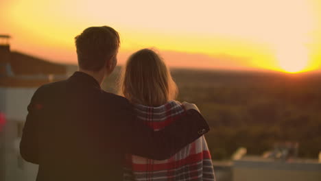 Liebende-Umarmen-Einen-Mann-Und-Ein-Mädchen-Und-Beobachten-Den-Sonnenuntergang-Bei-Einem-Glas-Wein,-Während-Sie-Auf-Dem-Dach-Eines-Gebäudes-Stehen.-Zeitlupenbild-Der-Beziehung-Eines-Jungen-Ehepaares.