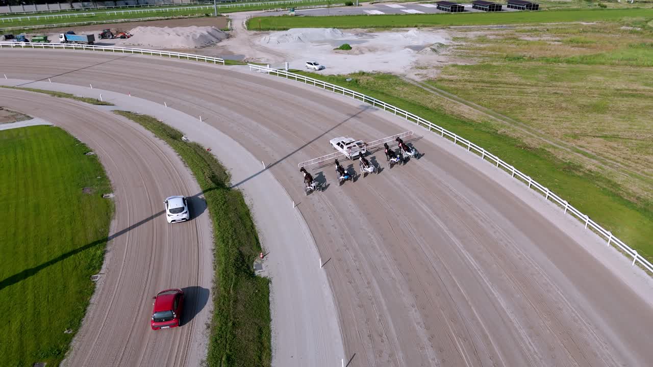 Aerial drone view of horses trotting on Milan racecourse during a competition, capturing dynamic motion, jockeys, track layout, and urban skyline in the background