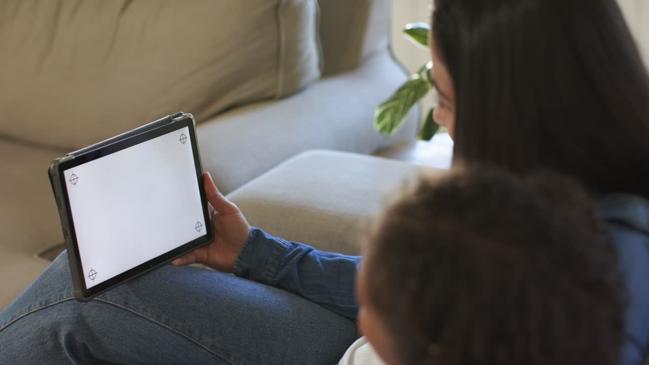 Mother reaching for tablet, turning it on and holding display toward child sitting on sofa cushion