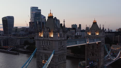 Elevated-footage-of-illuminated-top-walkway-and-decorated-towers-of-Tower-Bridge-after-sunset.-Skyscrapers-in-City-district-in-background.-London,-UK