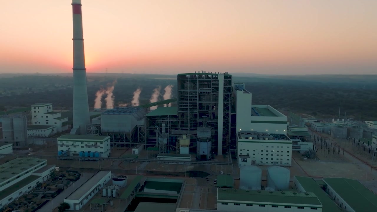 Drone flying toward a large industrial power plant during sunset with smokestack, steam columns and orbiting cinematic movement.