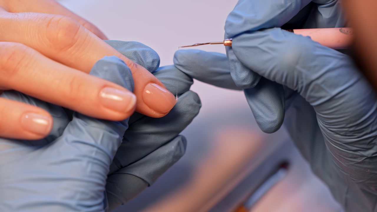 Elegant salon close-up of finished nude nails as a nail technician checks the manicure with a fine detail brush, highlighting glossy minimal beauty and careful hand styling.