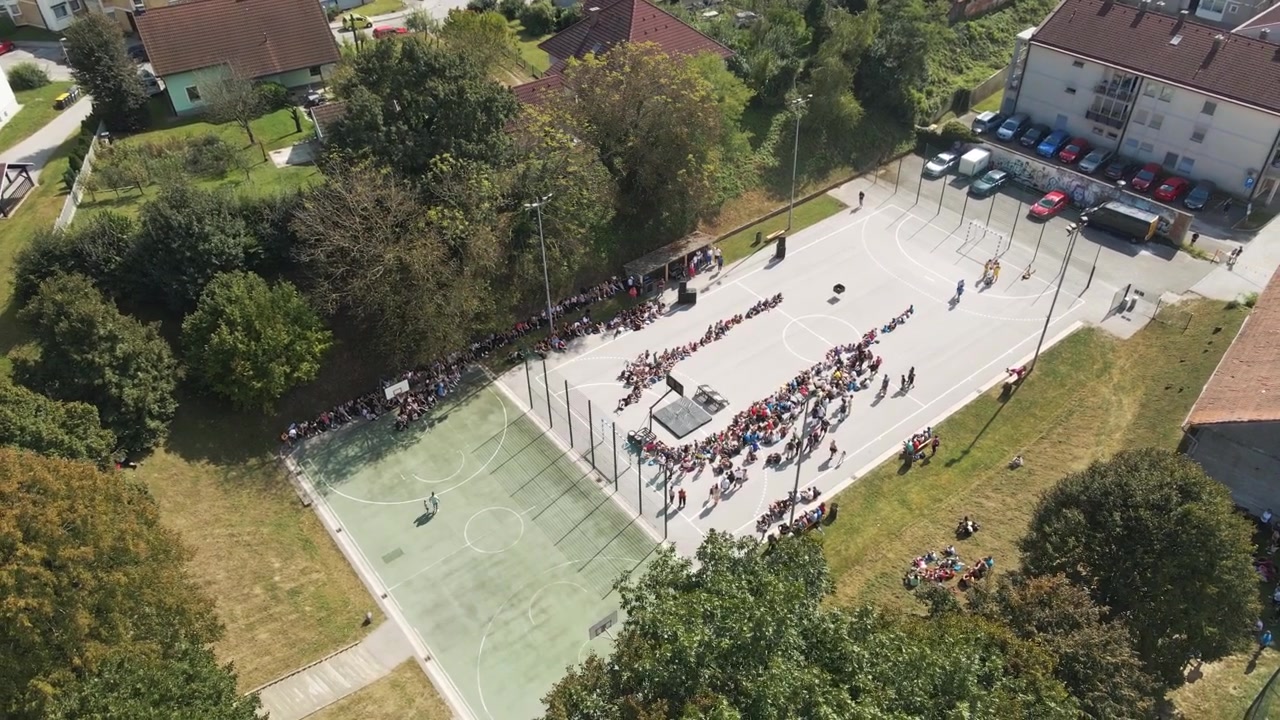 Video drone riprende una folla vivace riunita su un campo da basket all'aperto tra alberi verdi e case in una giornata di sole, creando un'atmosfera vibrante.