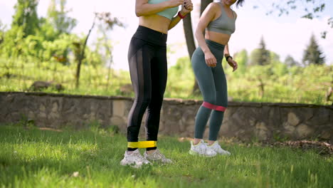 dos mujeres fitness haciendo pasos laterales y ejercicios de sentadillas con banda de resistencia en el parque