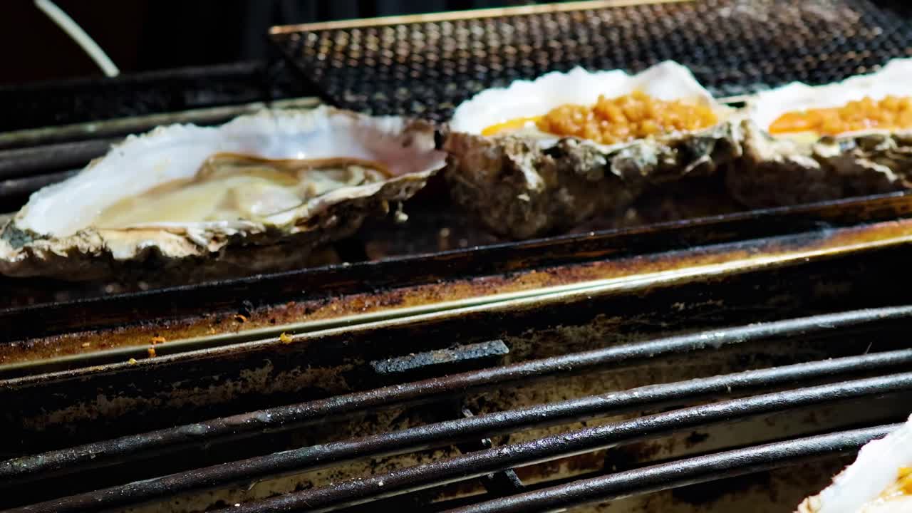 Detailed view of oysters being grilled, with tongs adding toppings on a hot grill.