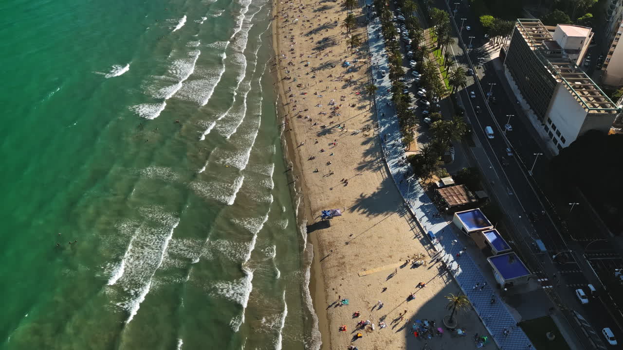 Aerial drone view of cars moving along the coastline with people relaxing on the beach in Alicante, Spain in daylight