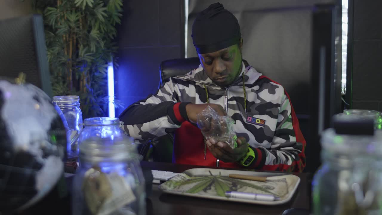 directamente a la vista de un hombre negro metiendo la mano en una bolsa de marihuana y quitando brotes que rompe en una bandeja de rollo de hojas de marihuana con un frasco de masón de dinero y un globo en el escritorio delante de él